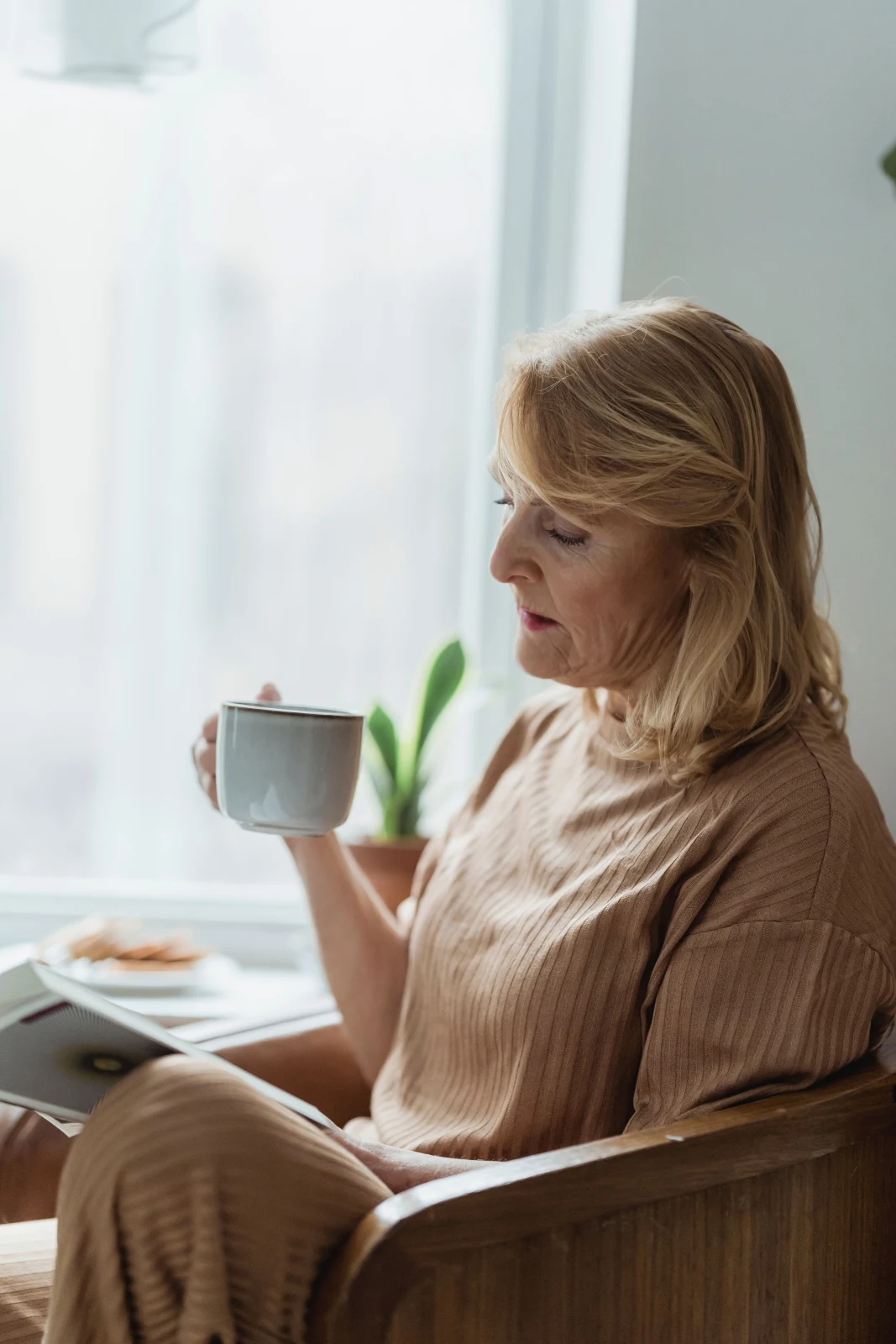 An older woman reads a book and holds a mug beside a bright window.